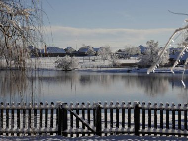 Kış sabahı çitleri karla kaplı taze kar yağmış bir göletin manzarası.