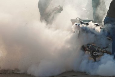 Duman tekerleklerin altından çıkıyor. Motosiklet tekerleği yakın plan. Lastiklerin asfalta sürtünmesinden dolayı duman. Sürücü motosiklet üzerindeki numarayı yapmaya hazırlanıyor. Yolda yanmış lastik..