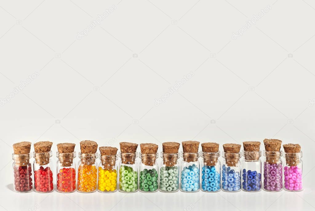 Red, yellow, blue, green, purple, pink and orange beads in glass jars on a white background. Beads in a transparent container with a wooden cork. All the colors of the rainbow are arranged in a row.