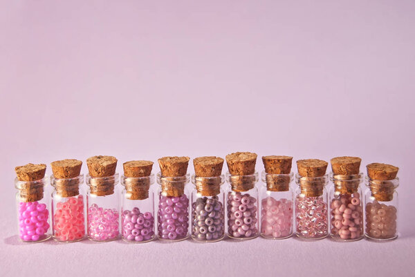 Beige, pink and yellow beads in glass jars on a bright purple background. Beads in a transparent container with a wooden cork. The concept of orderliness, balance and chaos.