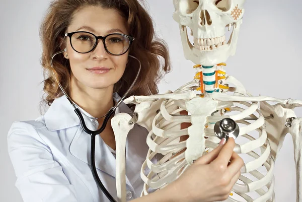 Woman in white medical gown. Doctor with skeleton layout on gray background. The doctor in the glasses listens to the ribs with a stethoscope.