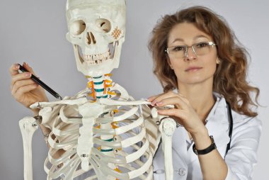 Tıbbi giysili kadın. İskelet yapısı olan bir doktor. Beyaz arka planda gözlüklü kız doktor. Doktorun asistanı kemiği işaret ediyor. Kas iskeleti sistemi hastalıkları üzerine çalışmalar.