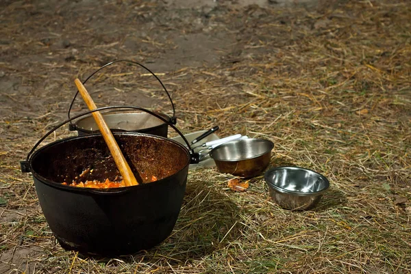 Food on a hike. Dinner is cooked on a fire. Food in a big black boiler. Metal bowl near the boiler.