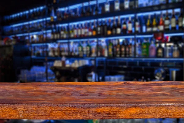 Wooden board on a background of bottles with alcohol. Old bar counter as layout for design. Workpiece for design. Empty place to advertise products. Blurred interior of the bar in the background.