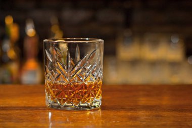 Bar tezgahında sarı kokteyl. Barın arka planında viski, rom ya da brendi. Ahşap bir tahtanın üzerinde içki bardağı.