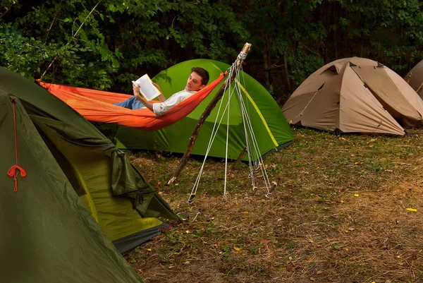 白いシャツの男が自然に本を読んでいる 男はテントの近くのオレンジのハンモックで休んでいる 森の近くの行にテントが配置されます — ストック写真