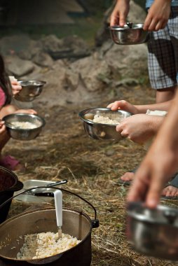 Çocuklar kamp gezisinde yemek yiyor. Kazanın içindeki yemek ateşte pişiyor. Kız ve oğlan maske takıyor. Çocuklar yiyecekleri tabağa koyarlar..