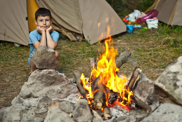 少年は火のそばに座っている 赤ん坊のシャープネス テントの背景に燃える炎 — ストック写真