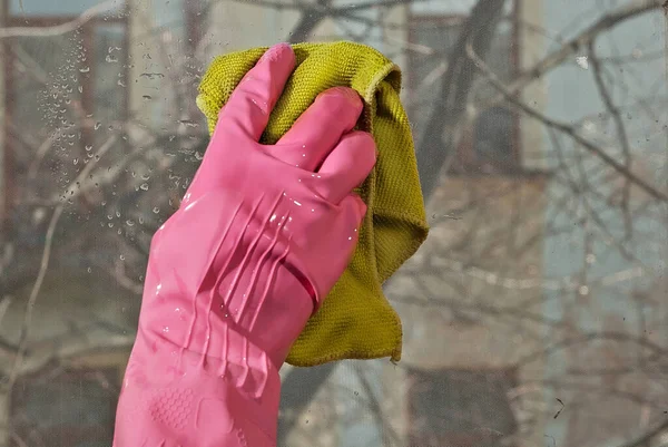Hand in pink rubber glove close up. The cleaner washes the window with a green cloth. Dirty dusty glass in sharpness. City street and buildings outside the window.