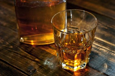 Şişenin yanında bir bardak alkol. Siyah ahşap masada eski bir viski şişesi. Karanlık arka planda içkiler bulanık.