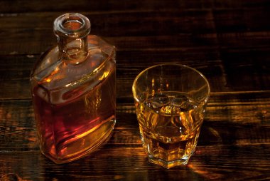 Şişenin yanında bir bardak alkol. Siyah ahşap masada eski bir viski şişesi. Karanlık arka planda içkiler bulanık.