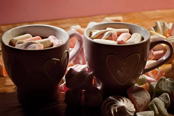 Lots of sweets near coffee mugs. Mugs on a pink background. The marshmallow dissolves into cappuccino.