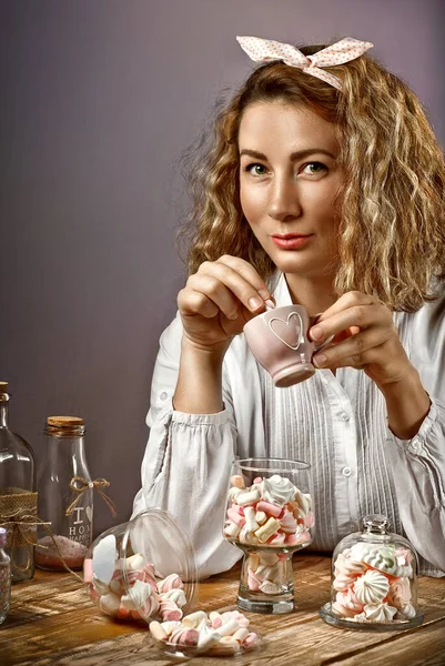 Girl at the table with sweets. Woman with a bow on her head sitting on a purple background. Lots of sweets in glassware on the table. Marshmallows, marmalade and bize in glasses on a wooden board.