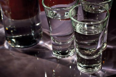 Şişenin yanında alkollü bardaklar. Mor arka planda şeffaf bir içecek..