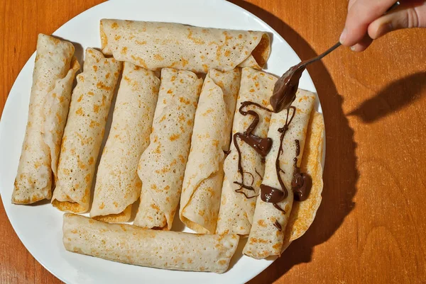 Pancakes are twisted into a tube and stuffed with cheese. Food in a white plate on a wooden table. Pancakes drizzled with chocolate syrup. Rolls with chocolate.