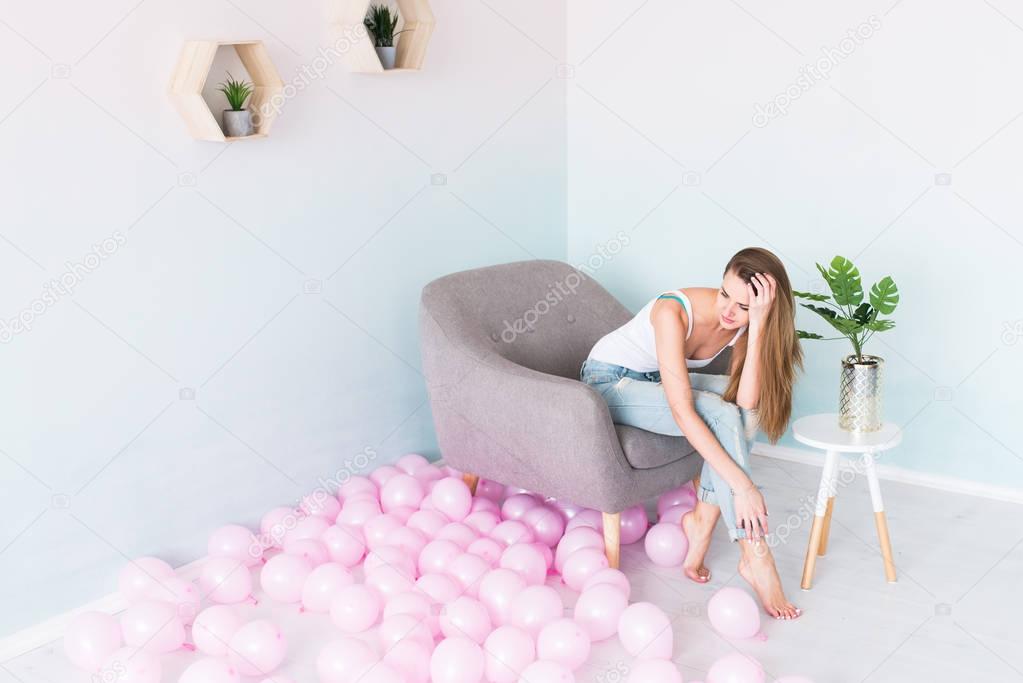 Young beautiful girl with long hair in white singlet and blue jeans with holes sitting in an armchair. Near a lot of pink balloons.