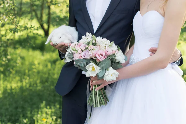 Novia sosteniendo en sus manos un delicado ramo de bodas con tulipanes blancos y rosados y rosas pequeñas rosadas. novio sosteniendo un blanco lindo conejo . — Foto de Stock