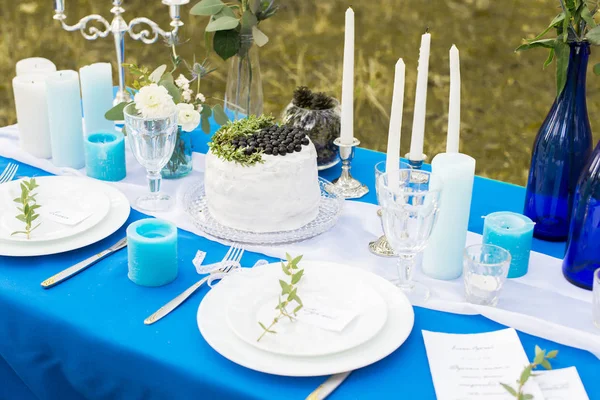 Mesa de banquete de casamento. Bolo com creme branco decorado com mirtilo e vegetação. Talheres com placas de stemware e velas em uma toalha de mesa azul . — Fotografia de Stock