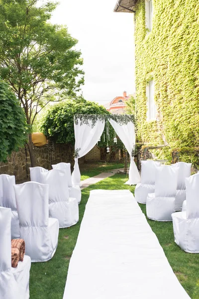 結婚式のアーチ白素材と緑がたくさん飾られています オープン空の下での結婚式のための美しいプラットフォーム 緑の草の新鮮な花で飾られた正方形のアーチ上の椅子 — ストック写真