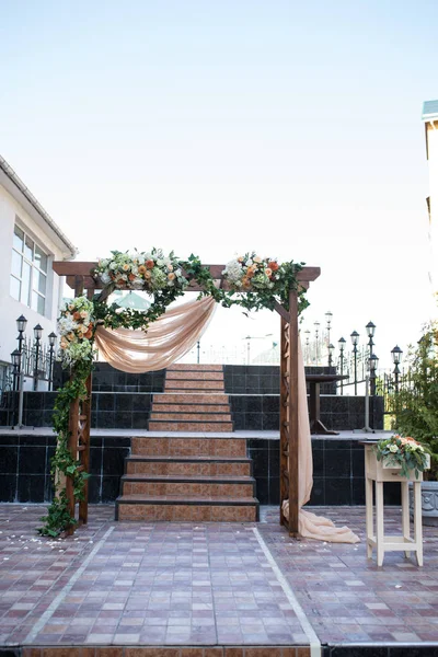 Rustic wedding arch. Wedding arch made of wooden square frame, beige cloth, white and orange flowers. Beautiful wedding ceremony outdoors.
