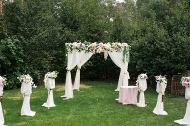 Açık havada düğün töreni için güzel platform: yeşil çimen, taze çiçeklerle süslenmiş kare kemer üzerinde ahşap sandalye