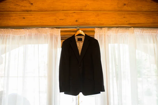 Beautiful black groom\'s jacket hanging on the window for morning wedding preparation. Grooms accessories