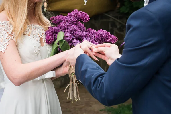 新郎は 花嫁の指に結婚指輪を置きます ライラックのブーケを持って花嫁 新郎新婦は 結婚式の日に指輪を交換します 結婚式のプロセス 結婚婚約 リング式の交換 — ストック写真
