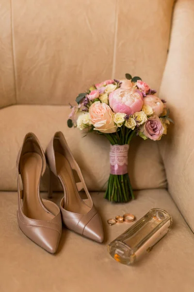 Bridal accessories: Wedding bouquet with pink peony, yellow, pink roses and greenery, elegant bridal beige shoes, golden rings, perfume. Wedding details