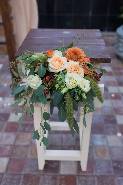 Rustic flower arrangement with beige roses, orange dahlia and a lot of greenery. Table decorated with flowers near the wedding arch. Wedding arrangement.