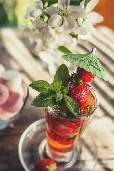 Detox drink made of ripe juicy strawberries and mint in a transparent glass Cup on the background of marshmallows and a bouquet of white flowers on a wooden table on a Sunny day. Selective focus.