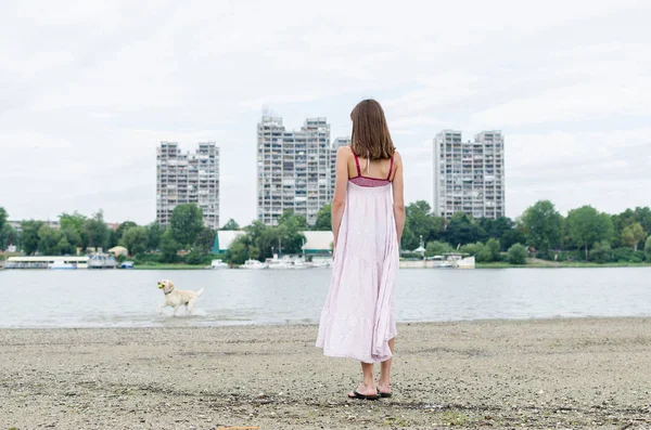 Fata de mers pe jos pe nisip lângă râu și oraș mare cu catelus — Fotografie, imagine de stoc