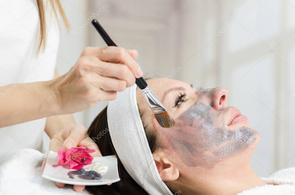 Cosmetologist applying grey mask on the client face.