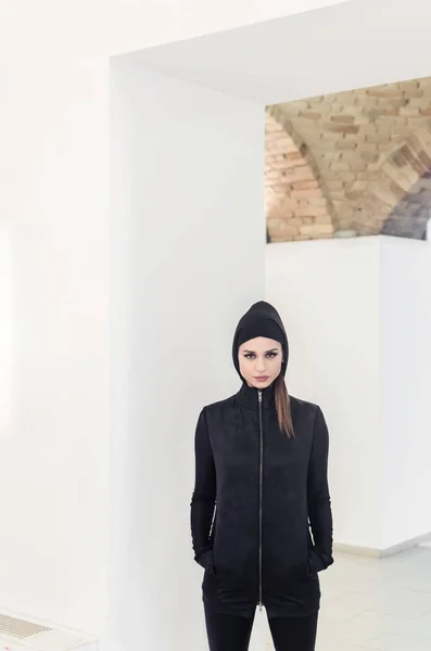 woman in black sportswear with hood on white wall background