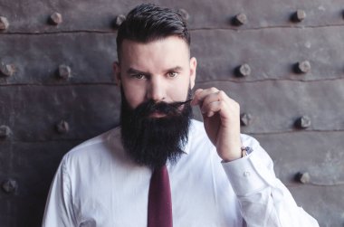 Handsome young bearded man in classic shirt holding tip of moustache and looking at camera clipart