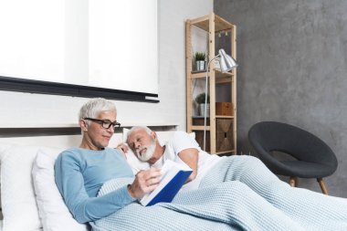 Modern evde, karı koca kitap okurken yatakta uzanan yaşlı çift uykuya daldı. Kopya alanı.