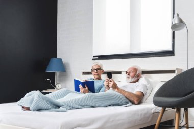 Kadın kitap okurken, koca akıllı telefondan mesaj gönderirken, yaşlı çift yatakta uzanıyor. Boşluğu kopyala