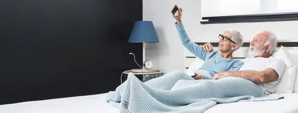 快乐的时刻 穿着睡衣躺在床上的一对快乐的老夫妇做了一个自拍 老婆拿着智能手机 — 图库照片