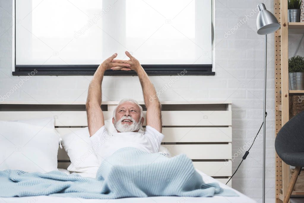 Portrait of a senior man in pajamas waking up in bed and stretching his arms