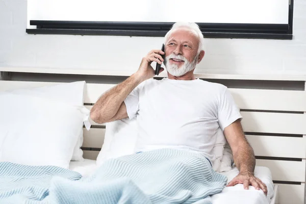 Senior man receive positive news over phone in bed in his room. Talking on the phone in pajamas after waking up with pleasure expression on face