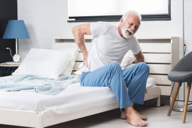 Senior man with white hair and beard alone sitting on bed and suffering from low back pain clipart