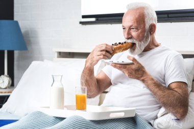 Happy senior man having breakfast in bed after waking up, eating chocolate croissant with milk and glass of orange juice clipart