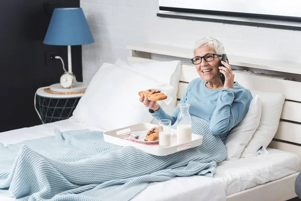 Senior woman eating breakfast in bed, in one hand chocolate croissant in another telephone and talk with her close friend