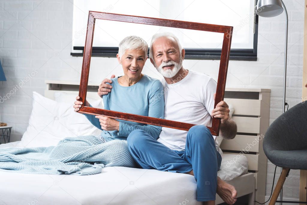 Portrait of senior couple in their pajamas on the bed with their hands hold a wooden frame while the photographer paints them