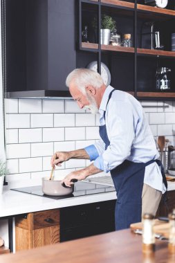 Kıdemli sakallı yakışıklı adamın fotoğrafı mutfakta duruyor ve önlükle öğle yemeği için sebze pişiriyor.