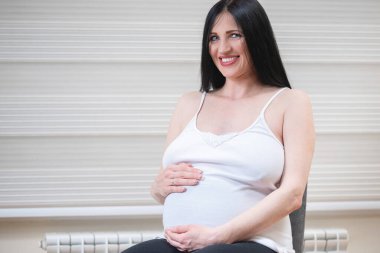 Kapalı fotoğraf, evde oturup karnını tutan hamile bir kadının portresi. Mutluluk kavramı, yeni hayat, yakında bebek kız doğacak.