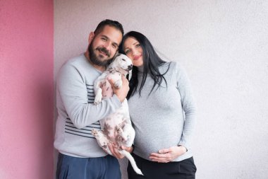 Geleceğin genç ebeveynleri ve güzel köpekleri bebek için duvarın önünde poz veriyorlar. Bebek gelişi, müstakbel ebeveynler, mutlu bir hayat, aile ferdi olarak evcil hayvan