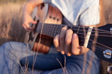 Kız gün batımında tarlada akustik gitar çalar. Parmaklar gitar panosunda. Doğa ve müzik. Yakın çekim..