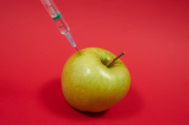 Zehir yeşil elmaya girer. Meyveli bir şırınga. Gmo konsept fotoğrafı. Kırmızı arka planda zehirlenmiş yemek. Yakın çekim..