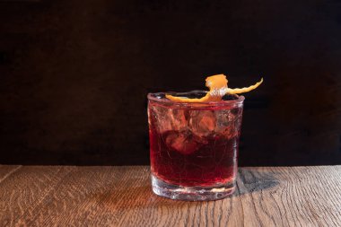 Kırık viski bardağı. Ahşap bir masada kırmızı alkol kokteyli. Buz ve portakal kabuğuyla servis edilmiş. Boulevardier veya Negroni. Loş ışıklı bar. Boşluğu kopyala.