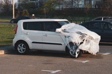 Park yerindeki bir kazadan sonra beyaz araba. Önden darbe. Plastik çarşafla kaplı kırık bir araba. Sigorta ödemesi bekleniyor.